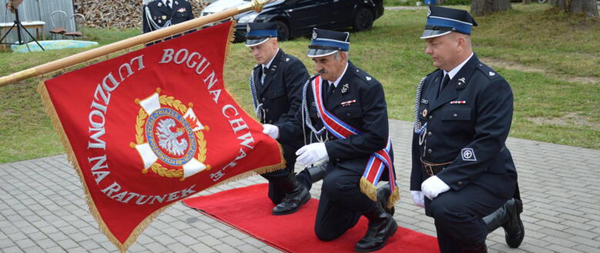 Zdjęcie przedstawia klęczący poczet sztandarowy na czerwonym dywanie przed wyciągniętym pochylonym sztandarem, w mundurach galowych. Na sztandarze znajduje się znak Związku OSP RP oraz napis Bogu na chwałę ludziom na ratunek.