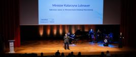 Widok na scenę na której stoi wiceminister Lubnauer i przemawia do mikrofonu. Na scenie też jest fortepian i zespół z instrumentami.