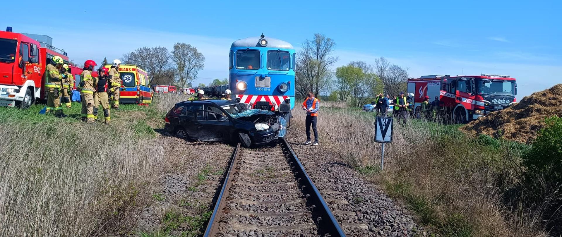 Zdjęcie zrobione z torowiska. Na zdjęciu widać cały teren akcji, lokomotywę która czołowo uderzyła w lewy bok samochodu osobowego. Po lewej i prawej stronie torowiska widać służby ratunkowe zadysponowane do zdarzenia.