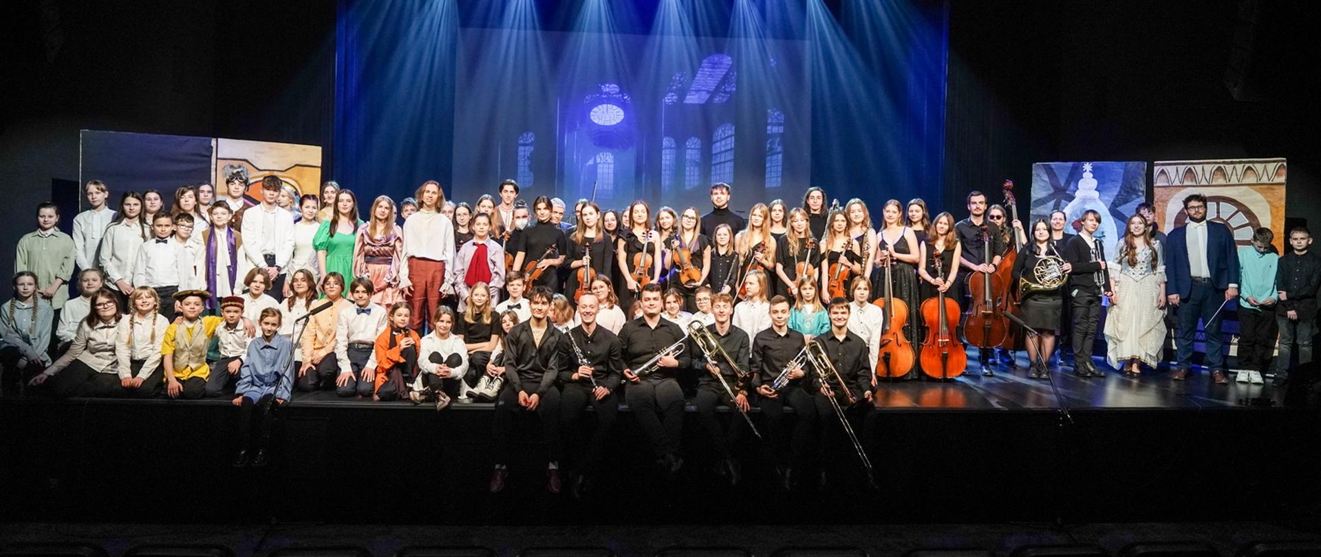 Zdjęcie przedstawia wykonawców spektaklu na scenie DK Oskard w Koninie, orkiestra ubrana na czarno trzyma instrumenty, pozostali wykonawcy w kolorowych strojach, dyrygent z batutą, w tle scenografia.