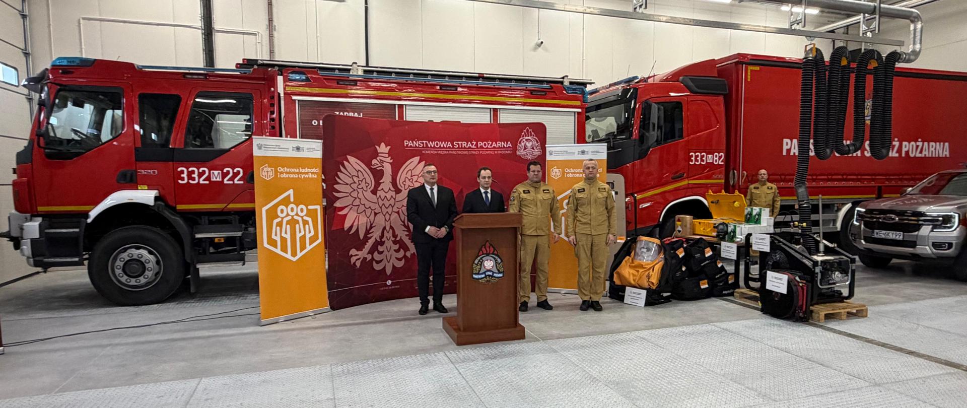 Zdjęcie zrobione we wnętrzu dużej hali garażowej. Od lewej kierownik delegatury Mazowieckiego Urzędu Wojewódzkiego w Radomiu Pan Rafał Molga i Wicewojewoda Mazowiecki Pan Patryk Fajdek w czarnych garniturach oraz Mazowiecki Komendant Wojewódzki PSP nadbryg. Artur Gonera i Komendant Miejski PSP w Radomiu st. bryg. Łukasz Szymański w piaskowych mundurach służbowych. Za nimi widoczna czerwona ścianka z białym napisem „PAŃSTWOWA STRAŻ POZARNA | „KOMENDA MIEJSKA PAŃSTWOWEJ STRAŻY POŻARNEJ W RADOMIU” oraz żółte roll-upy z białym napisem „Ochrona ludności i obrona cywilna”. Za ściankami widoczne dwa czerwone wozy strażackie, a po prawej stronie zdjęcia strażak PSP w piaskowym mundurze służbowym i srebrny samochód. 