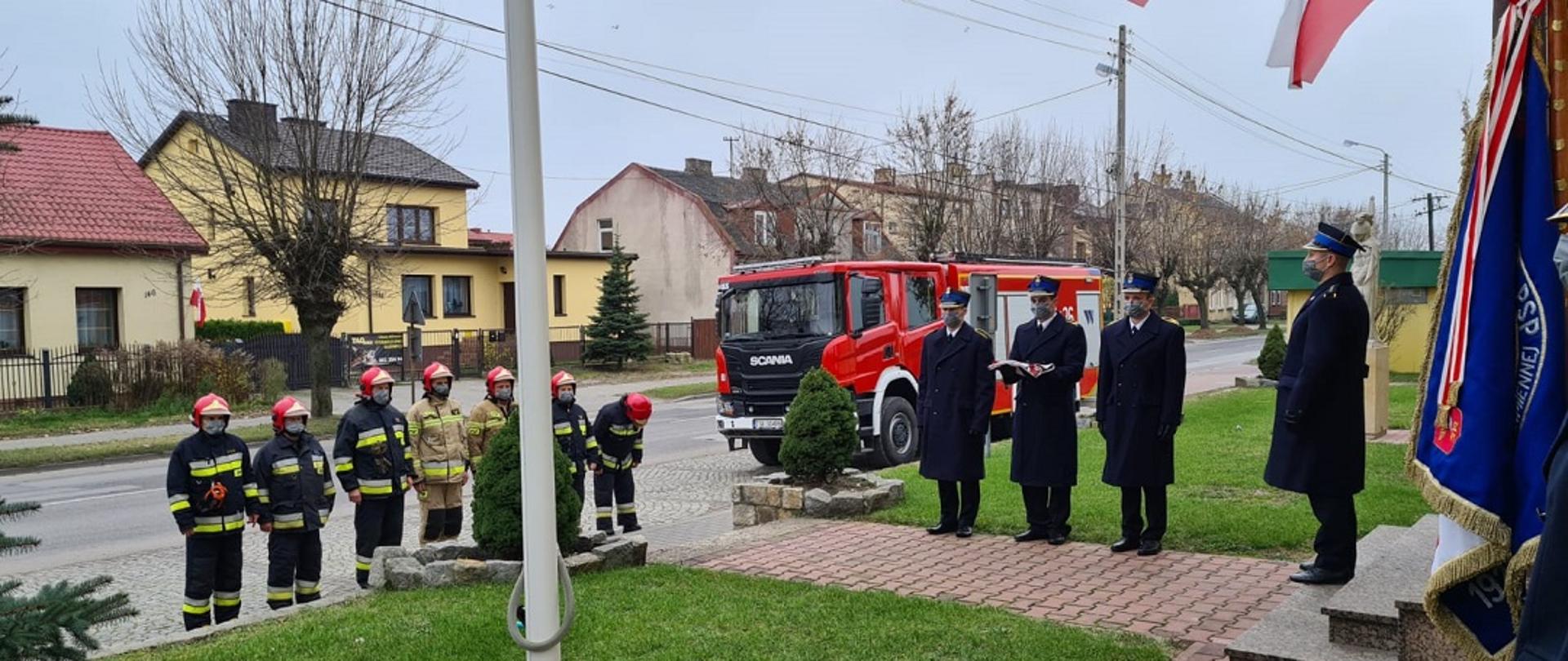 Poczet flagowy oraz ratownicy z JRG Skarżysko-Kamienna podczas uroczystego apelu z okazji Święta Niepodległości. Poczet flagowy składający się z trzech funkcjonariuszy stoi na tle samochodu gaśniczego przed budynkiem KP PSP w Skarżysku-Kamiennej. Na schodach przed wejściem do budynku stoi dowódca zmainy. Wszyscy ubrani są na galowo. Na wprost dowódcy zmiany w ubraniach bojowych w szeregu stoją ratownicy pełniący służbę.