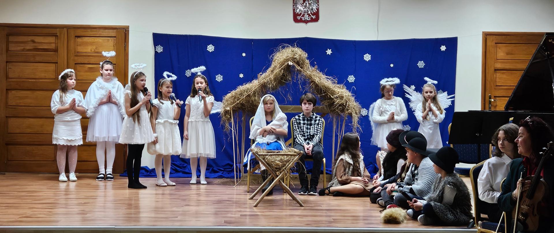 Fotografia przedstawia grupę młodych aktorów z klasy III c. 6 ,stojących na scenie Auli PSM I st. w Grajewie. Z tyłu stoją anioły na tle niebieskiej scenografii z gwiazdami imitującej niebo, obok stajenka, a przed nią siedzą Józef i Maryja trzymająca w ramionach niemowlę, przed nimi stoi żłóbek. Obok po prawej stronie siedzą pastuszkowie i pastereczki.