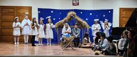 Fotografia przedstawia grupę młodych aktorów z klasy III c. 6 ,stojących na scenie Auli PSM I st. w Grajewie. Z tyłu stoją anioły na tle niebieskiej scenografii z gwiazdami imitującej niebo, obok stajenka, a przed nią siedzą Józef i Maryja trzymająca w ramionach niemowlę, przed nimi stoi żłóbek. Obok po prawej stronie siedzą pastuszkowie i pastereczki.