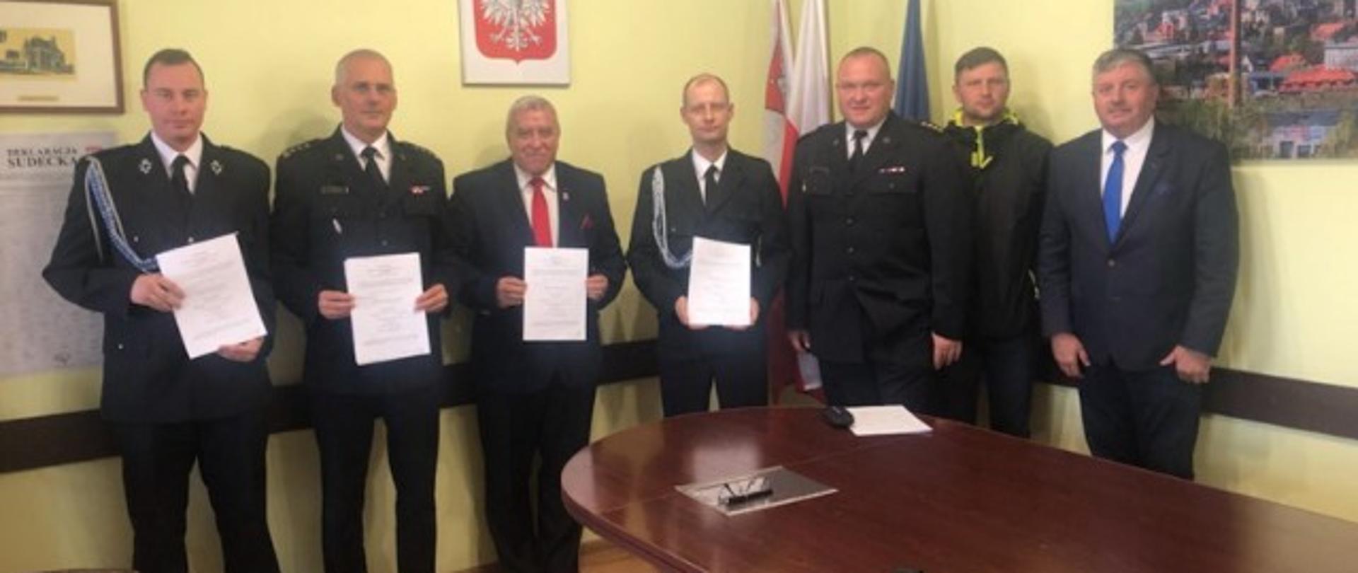 Panowie w mundurach wyjściowych oraz garniturach stoją przy ścianie i trzymają przed sobą podpisane dokumenty.