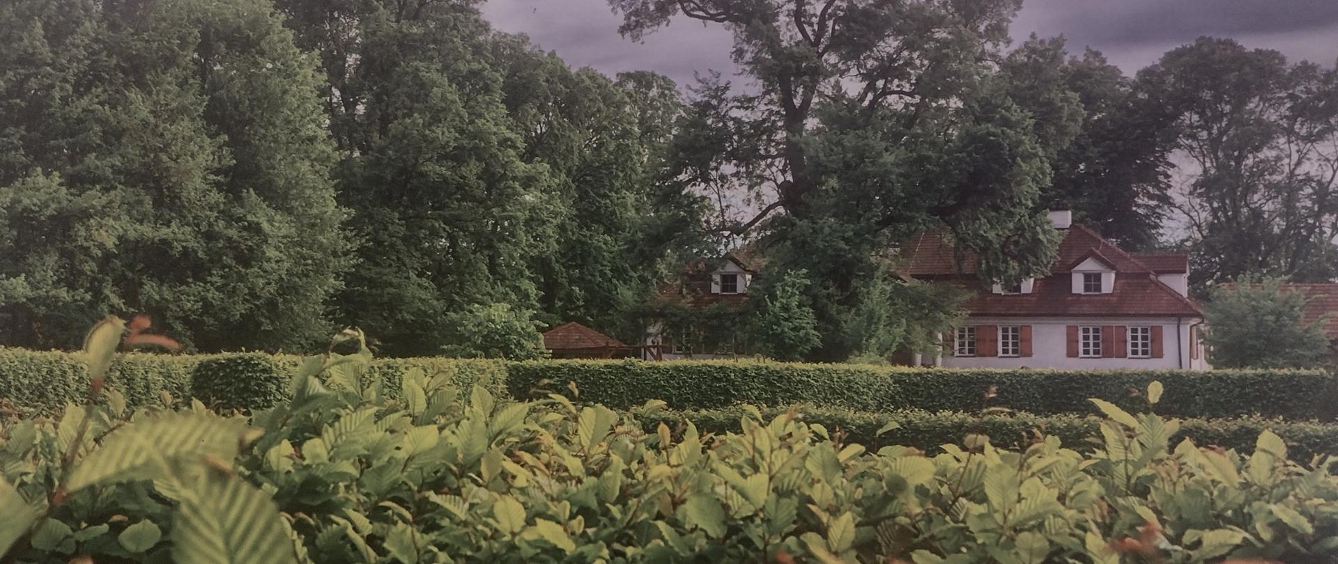Zdjęcie przedstawiające dworek wśród zieleni drzew i krzewów.