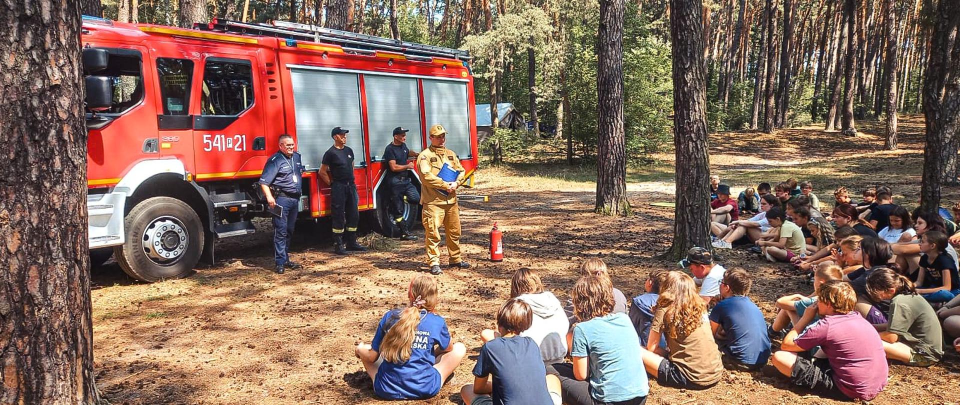Strażacy wspólnie z policjantami omawiają zasady bezpieczeństwa podczas obozu harcerskiego.
