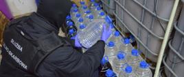 Funkcjonariusz Służby Celno-Skarbowej w kominiarce kuca przy ujawnionym nielegalnym alkoholu w plastikowych butelkach.