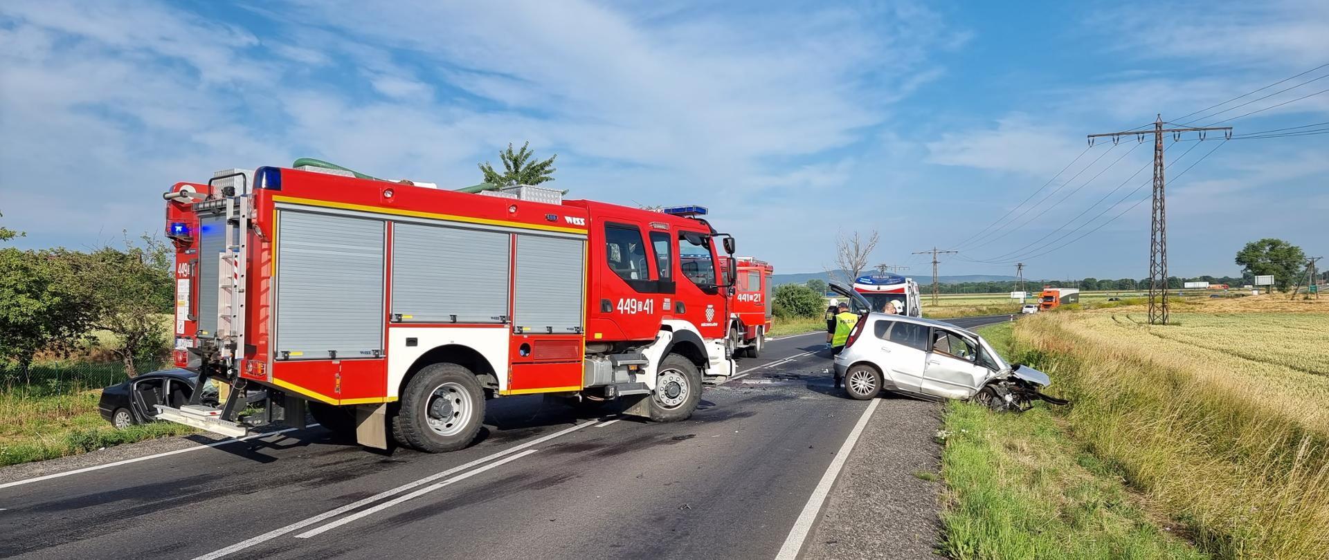 Obraz przedstawia wrak pojazdu po wypadku. Pojazd stoi częściowo na poboczu i przydrożnym rowie. Obok pojazd pożarniczy. W tle karetka pogotowia ratunkowego.