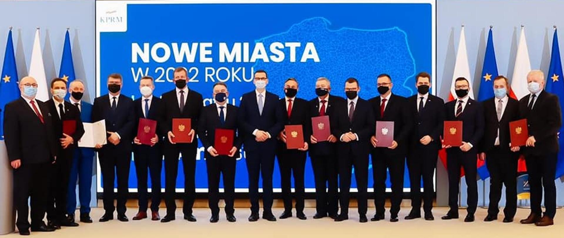 Na zdjęciu znajduje się 16 elegancko ubranych mężczyzn. Pozują oni do zdjęcia, stojąc na podeście. W tle granatowa grafika przygotowana przez Kancelarię Premiera z napisem "NOWE MIASTA W 2022 ROKU". 