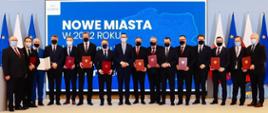 Na zdjęciu znajduje się 16 elegancko ubranych mężczyzn. Pozują oni do zdjęcia, stojąc na podeście. W tle granatowa grafika przygotowana przez Kancelarię Premiera z napisem "NOWE MIASTA W 2022 ROKU".