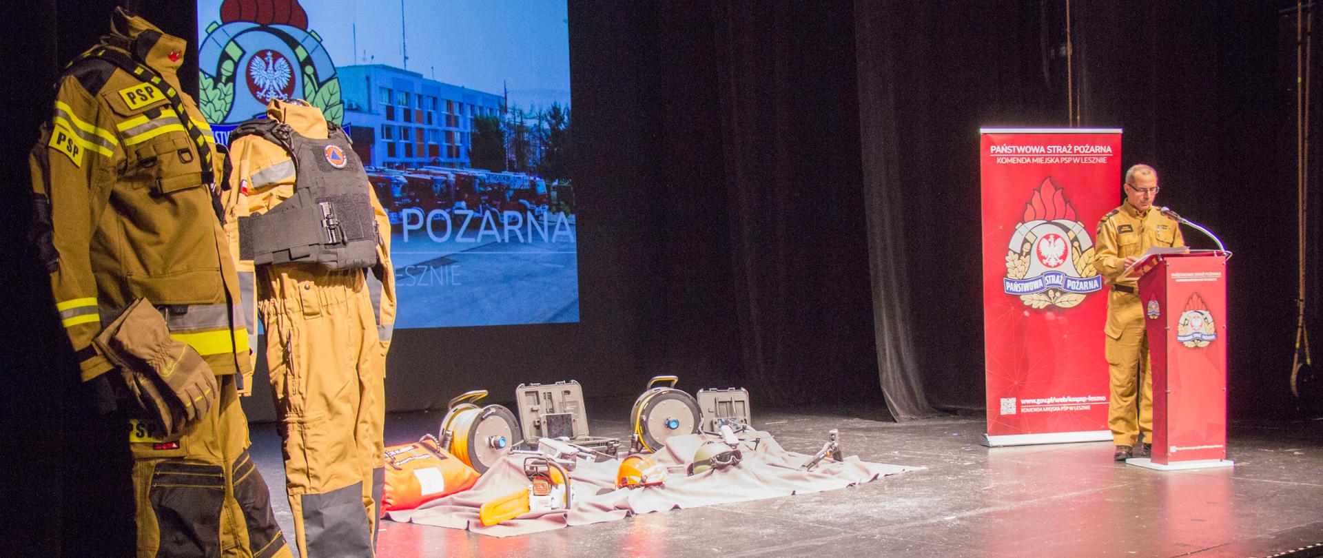 Z lewej strony widać ubrania specjalne na manekinach, w centralnej części znajduje się scena na której ułożone są sprzęty ratownicze. Z prawej strony strażak przemawia przy mównicy z logiem PSP.