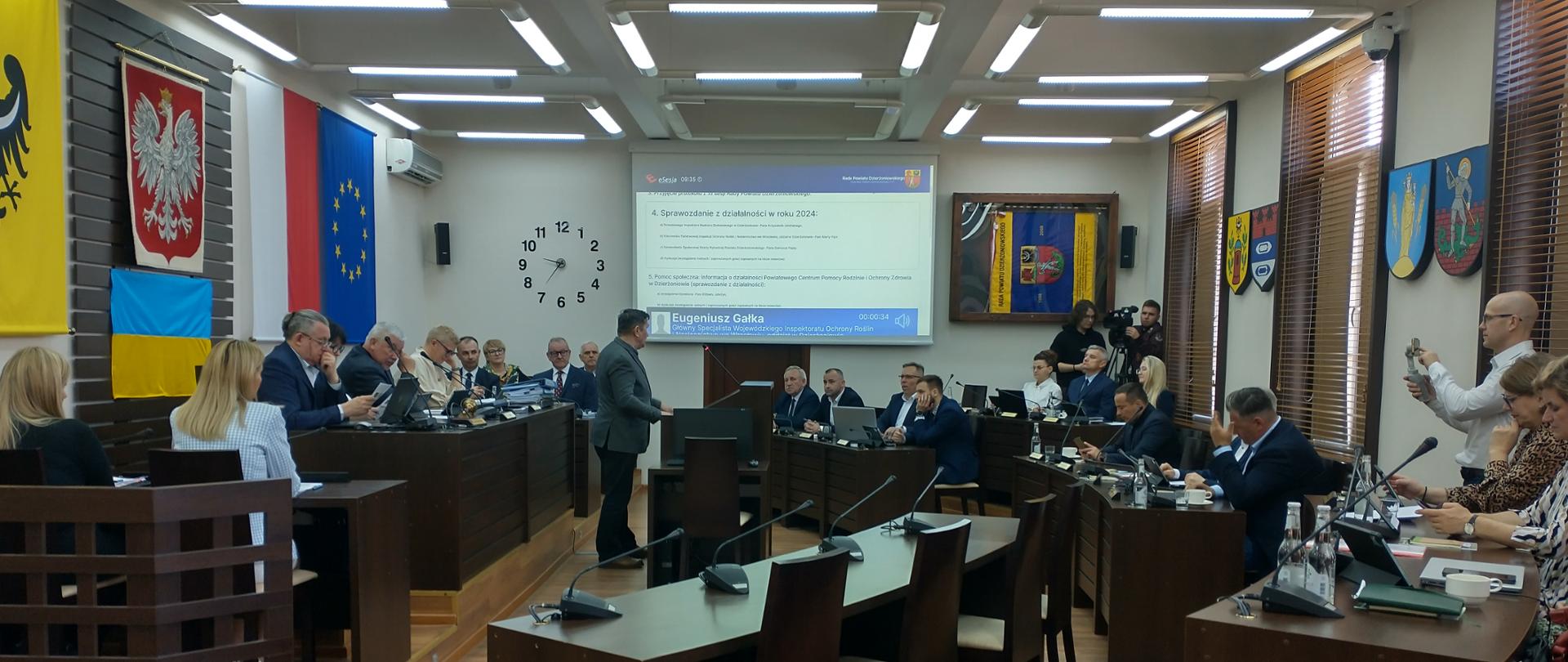 Inspektor Oddziału w Dzierżoniowie przedstawiający prezentację w sali na której w półkolach ustawione są stoły przy których siedzą uczestnicy spotkania. Na ścianie flagi Polski, UE, Powiatu Wrocławskiego.