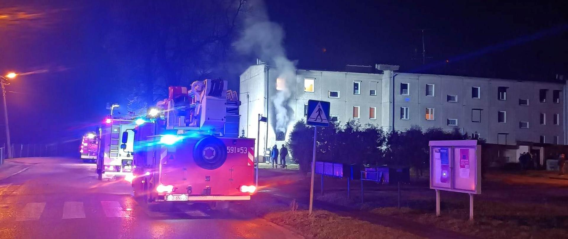 Po prawej widać budynek wielorodzinny z którego wydobywa się dym, po lewej samochody strażackie