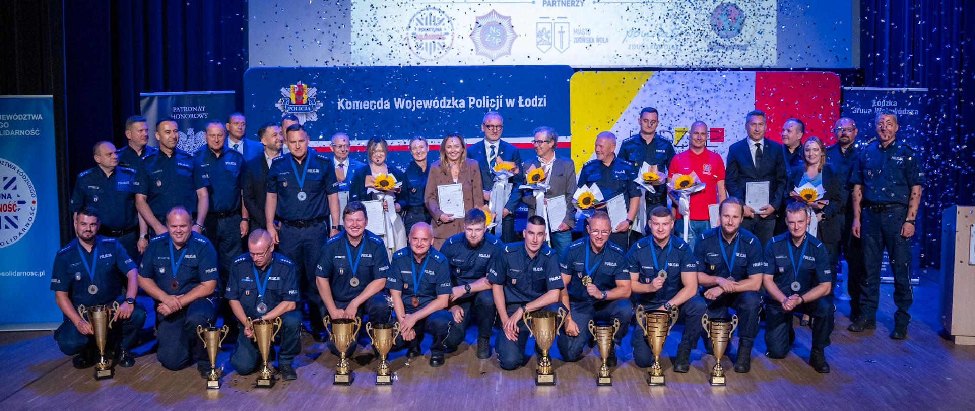 Zdjęcie grupowe uczestników gali konkursowej - na pierwszym planie znajdują się policjanci z pucharami, w drugim rzędzie - nagrodzeni wspólnie z przedstawicielami władzy 