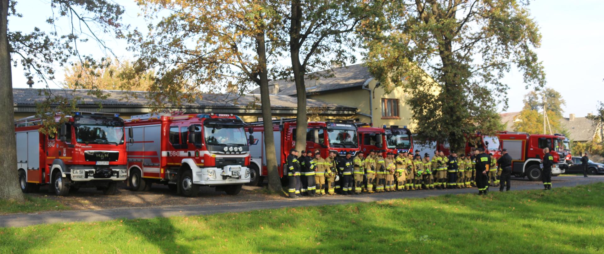Odprawa strażaków przed rozpoczęciem ćwiczeń. Na drugim planie wozy strażackie przewidziane do ćwiczeń.