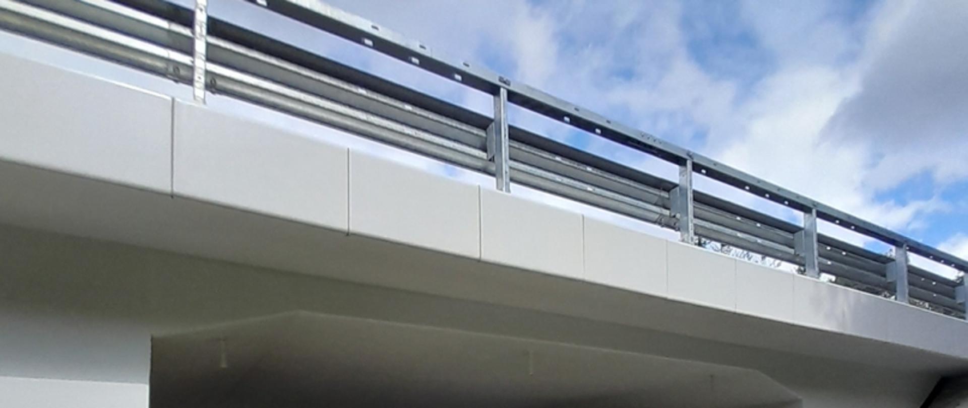 Zdjęcie przedstawia fragment mostu lub wiaduktu z metalowymi elementami i widokiem na niebo w tle.