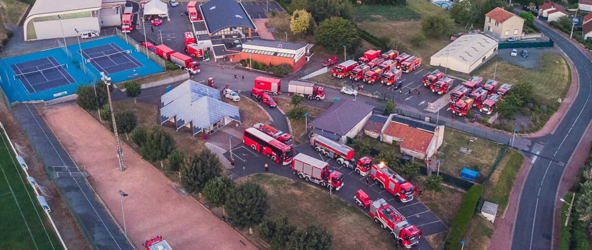 Widok z lotu ptaka miejsca postoju polskich strażaków. Widocznych jest kilkadziesiąt samochodów pożarniczych zaparkowanych w miejscu postoju