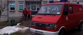 Na pierwszym palnie stoi zaparkowany samochód pożarniczy przed przychodnią. W tle widać strażaka-ochotnika, który pomaga wejść do przychodni pacjentce