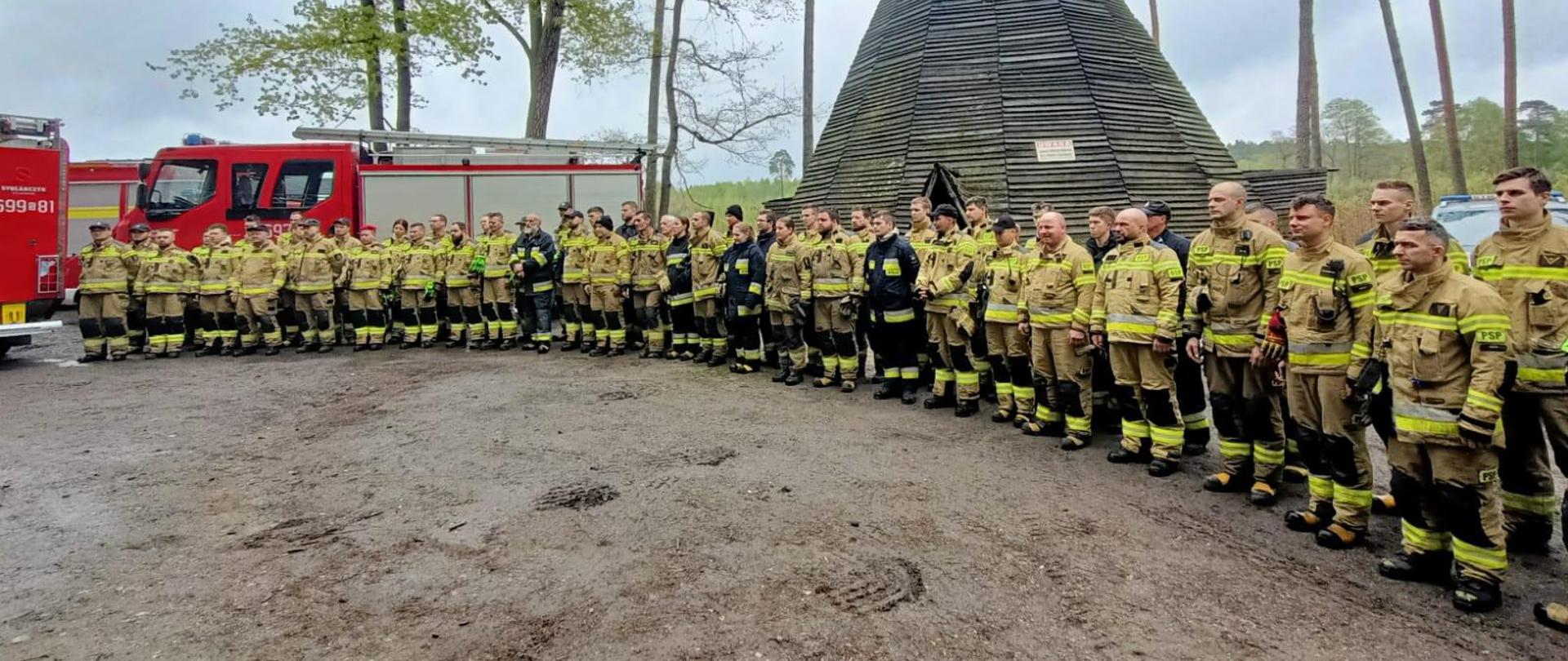 Strażcy stoją w półkolu na placu na podumowaniu ćiwczeń. Za nimi stożkowaty budynek - szałas. i samochody pożarnicze. W oddali drzewa.