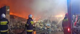 Na zdjęciu w porze nocnej widać palące się śmieci, głownie tworzywa sztuczne. Na zdjęciu panuje silne zadymienie. Na pierwszym palnie po prawej stronie widać od tyłu samochód gaśniczy z którego podawana jest woda. Po lewej stronie natomiast dwóch strażaków przelewa przegarnięte odpady z węża strażackiego. Przy samochodzie gaśniczych stoi kierowca.