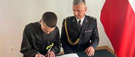 Nowoprzyjęty strazak podpisuje akt ślubowania obok siedzi Komendant Powiatowy