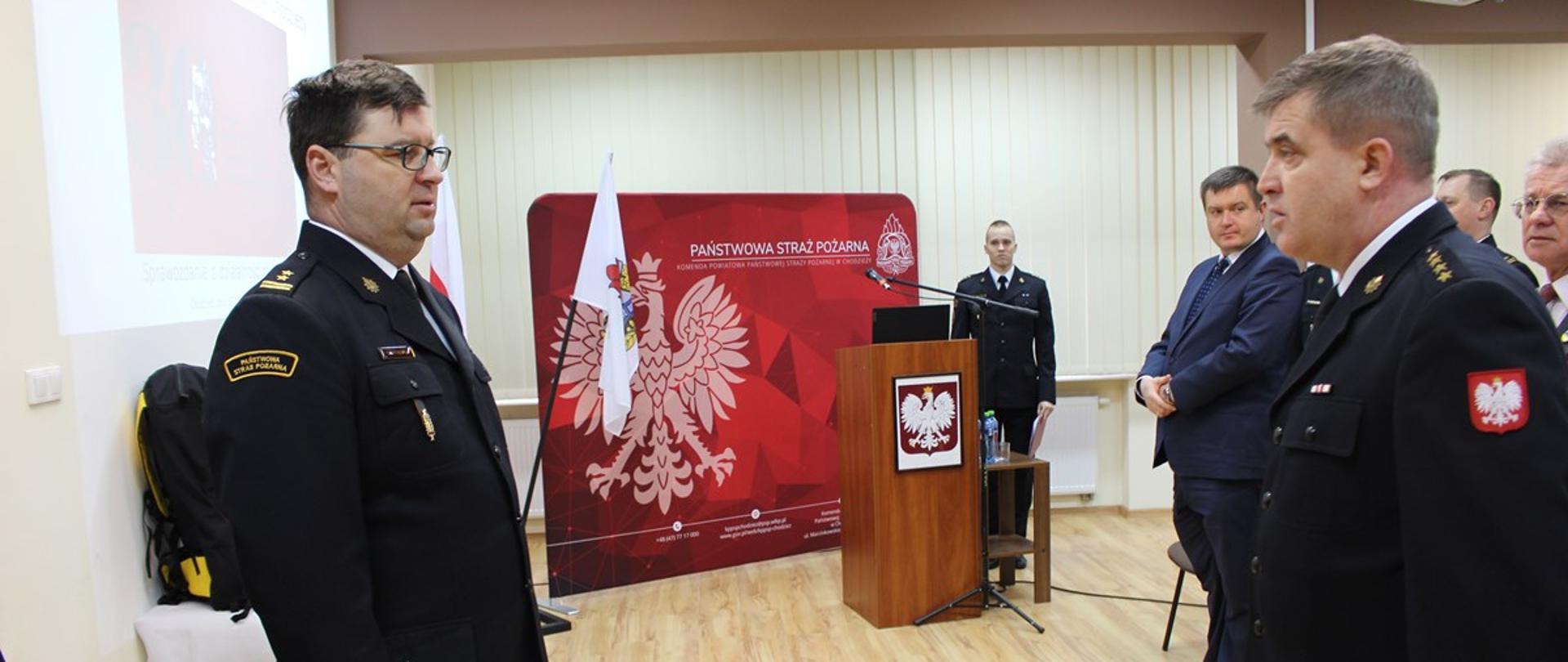 składanie meldunku strażakowi przez innego strażaka, w tle mównica z mikrofonem a za nią czerwony baner z orłem i biała flaga.