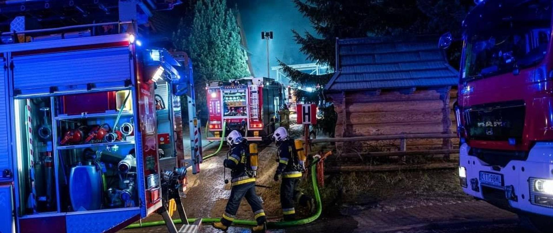 Zdjęcie przedstawia działania straży pożarnej podczas pożaru w Kościelisku 9 grudnia 2020 roku. Na zdjęciu widać wozy strażackie, strażaków w sprzęcie ochrony dróg oddechowych. W tle widać unoszący się dym nad budynkami. Tło ciemne z uwagi na wczesne poranne godzin y pożaru.