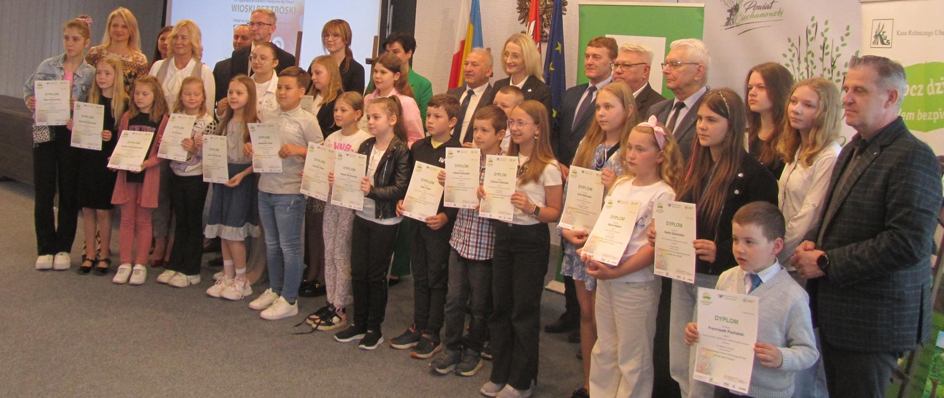 W reprezentacyjnej sali urzędu, na tle monitora z nazwą konkursu, flag i bannerów z logo KRUS pozuje do zdjęcia 21 nagrodzonych dzieci stojących w dwóch rzędach przed grupą kobiet i mężczyzn stojących za nimi. Młodzi laureaci eksponują swoje dyplomy.