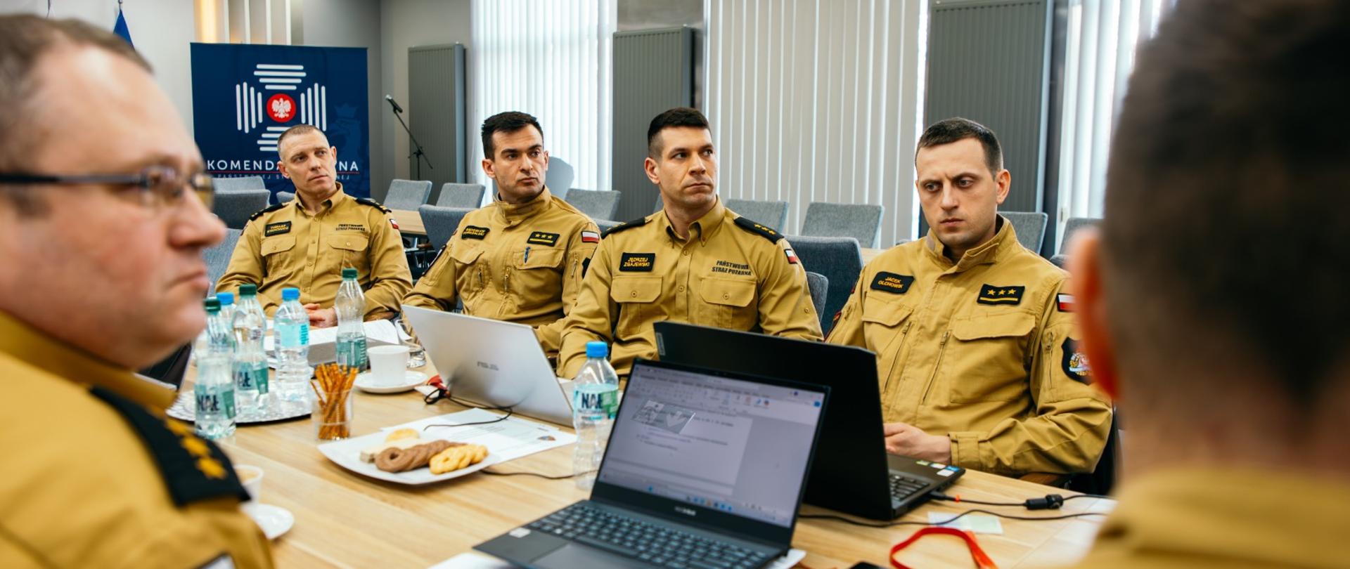 Kilku strażaków w mundurach służbowych siedzą z obu stron dużego stołu. Na stole laptopy, notatki, woda i herbatniki. W tle baner firmowy.