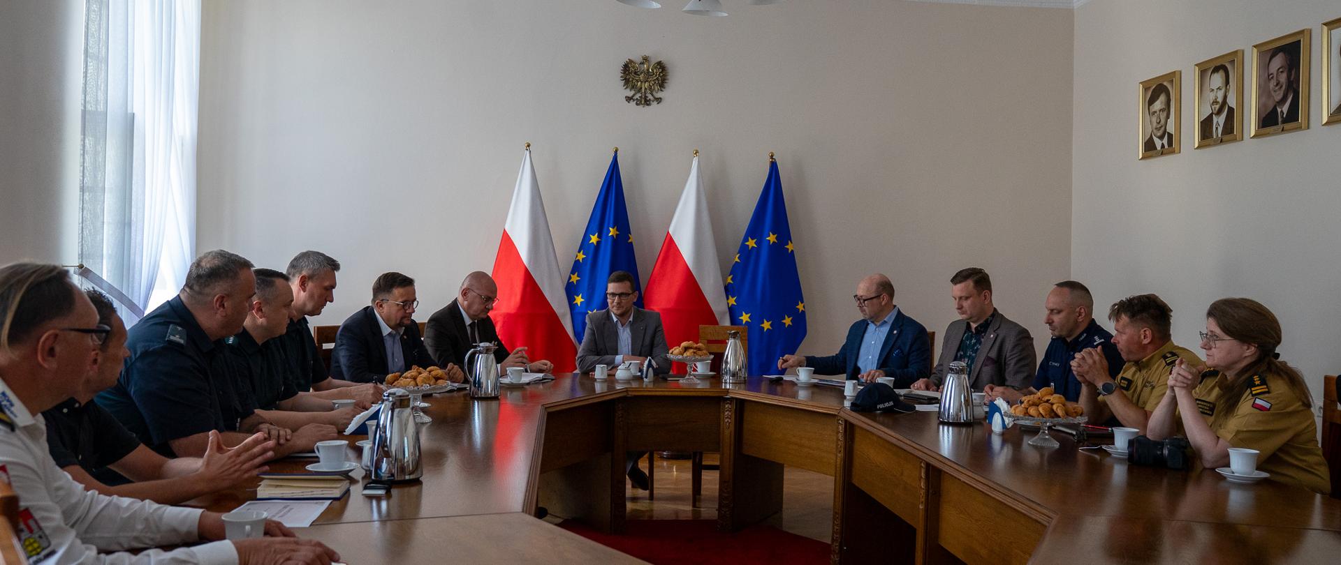 Przy okrągłym stole wielu obradujących, po prawej stronie widać dwóch strażaków, za stołem, po środku cztery flagi, naprzemiennie - narodowa biało-czerwona i niebieska w gwiazdy w okręgu - unijna.