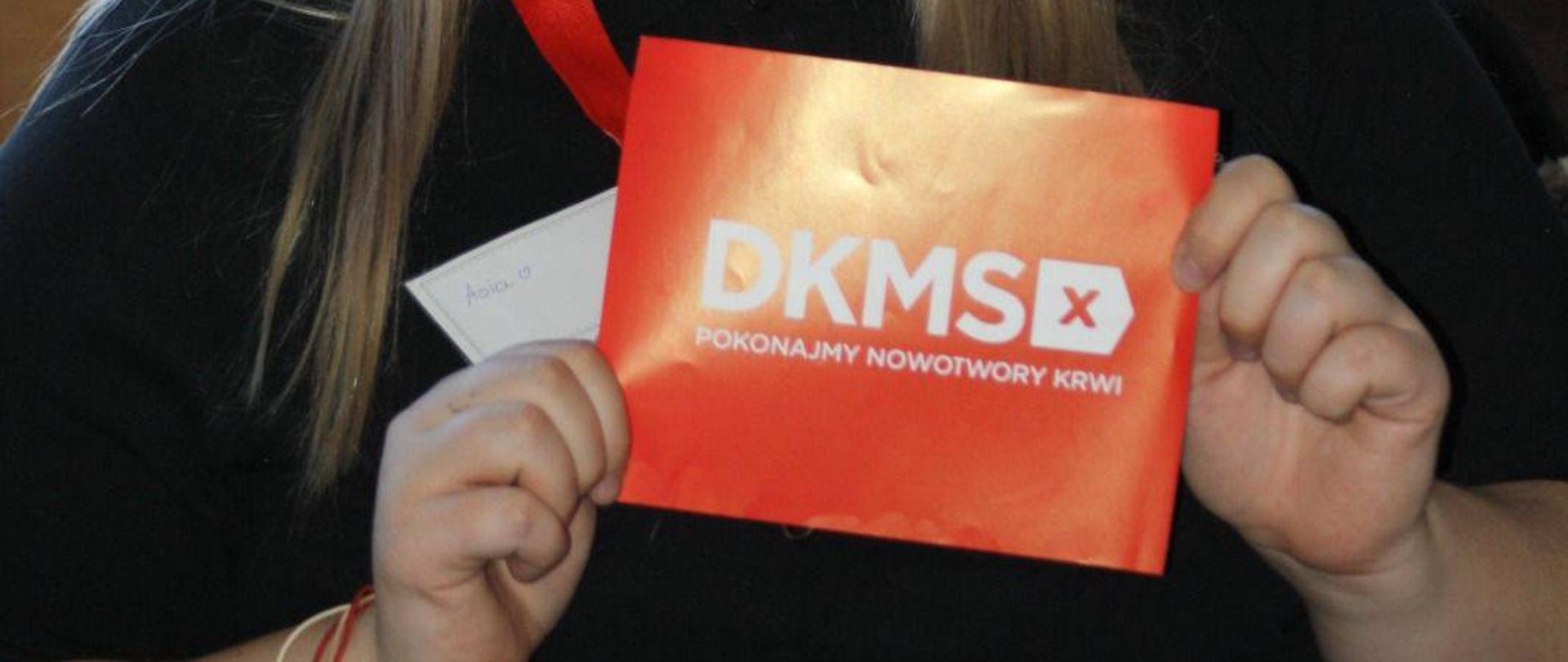 Uśmiechnięta młoda kobieta o jasnych włosach, trzymająca przed sobą czerwoną tabliczkę z białym napisem „DKMS. Pokonajmy nowotwory krwi”. W tle widać gazetkę ścienną z motywami czerwonych serc i kwiatów oraz materiały informacyjne dotyczące pobierania komórek macierzystych.