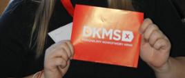 czerwona tabliczka z białym napisem „DKMS. Pokonajmy nowotwory krwi”. W tle widać gazetkę ścienną z motywami czerwonych serc i kwiatów oraz materiały informacyjne dotyczące pobierania komórek macierzystych.