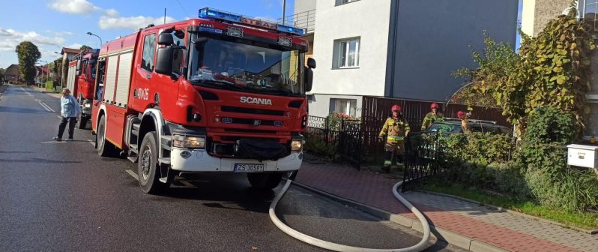Na pierwszym planie dwa wozy straży pożarnej stojące wzdłuż chodnika. Do jednego z aut podłączona jest linia wężowa. Na drugim planie strażacy w piaskowych mundurach prowadzący działania ratownicze