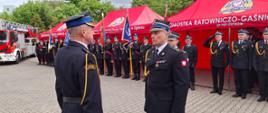 Zdjęcie przedstawia Zastępcę Dowódcy Jednostki Ratowniczo-Gaśniczej w Zgierzu podczas składania meldunku Zastępcy Łódzkiego Komendanta Wojewódzkiego w Łodzi. Na drugim planie widać poczty sztandarowe straży pożarnych.
