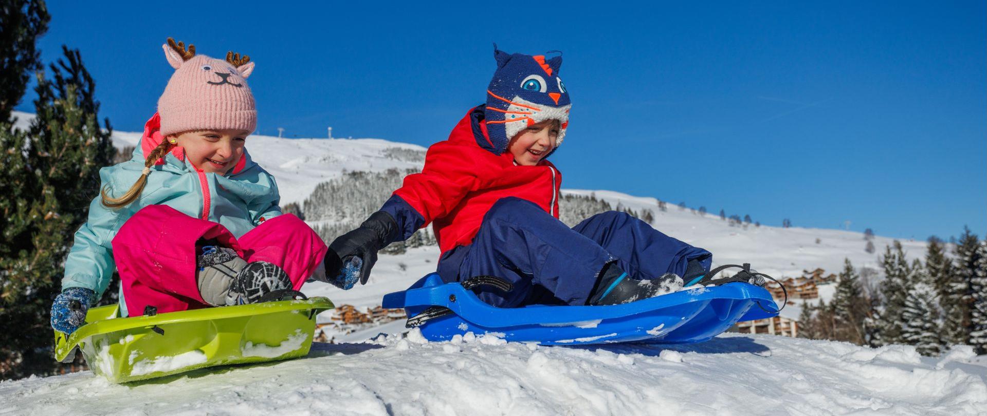 Na zdjęciu widać dwoje dzieci bawiących się na śniegu, siedzących na saneczkach na szczycie niewielkiego wzniesienia. Jedno z nich ma zielone sanki i zimowy strój w jasnych, pastelowych kolorach oraz różową czapkę z motywem zwierzątka. Drugie siedzi na niebieskich sankach, ubrane w czerwoną kurtkę, granatowe spodnie i charakterystyczną czapkę z motywem kota.
W tle rozciąga się zimowy krajobraz: ośnieżone stoki, drzewa pokryte śniegiem oraz czyste, intensywnie niebieskie niebo. Scena sprawia wrażenie pełnej radości i zimowej zabawy na świeżym powietrzu.