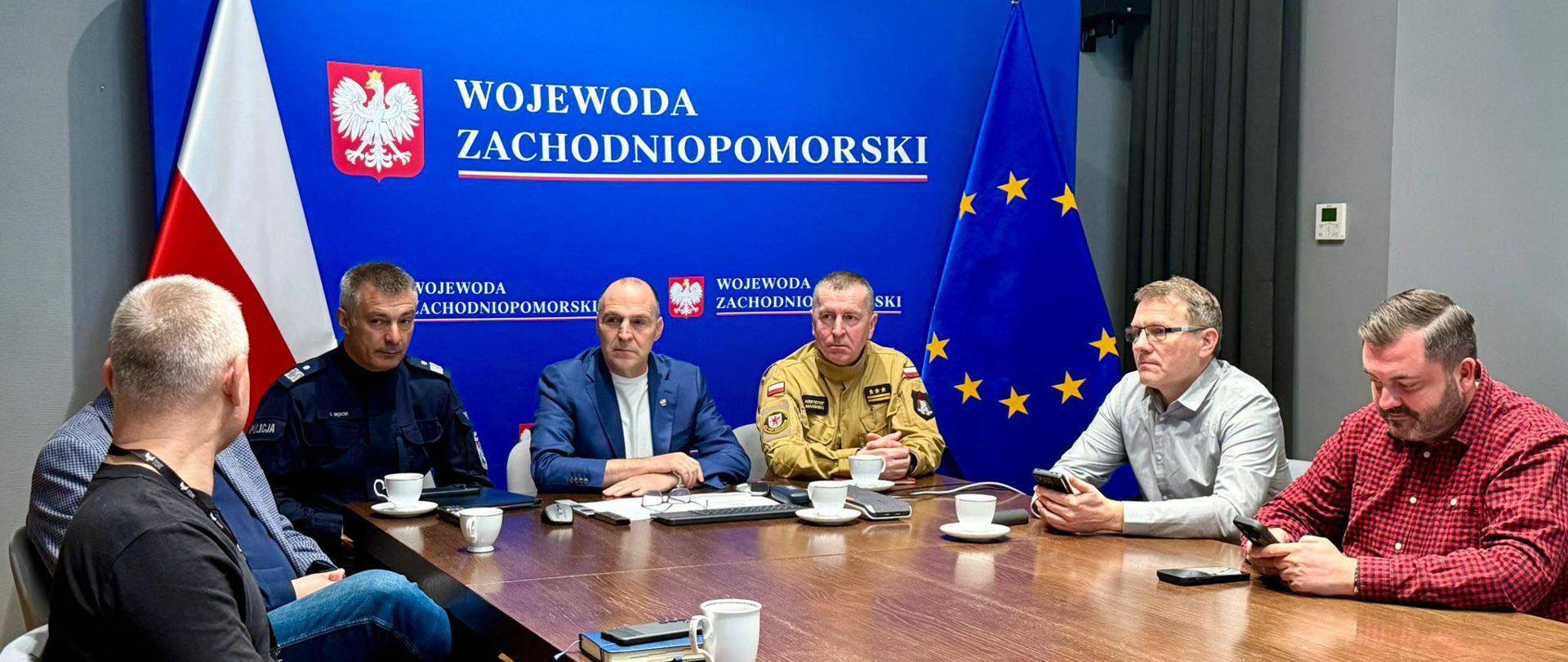 W pomieszczeniu konferencyjnym siedem osób bierze udział w spotkaniu przy dużym, prostokątnym drewnianym stole. Na ścianie za nimi znajduje się niebieskie tło z napisem „Wojewoda Zachodniopomorski” oraz herbem Polski. Po lewej stronie stoi flaga Polski (biało-czerwona), a po prawej flaga Unii Europejskiej (niebieska z żółtymi gwiazdami).
Przy stole siedzą głównie mężczyźni w średnim wieku. Dwóch z nich ma na sobie mundury służbowe – jeden w ciemnym stroju policyjnym, drugi w jasnym, przypominającym mundur służb ratowniczych. Pozostali ubrani są w stroje cywilne: marynarki, koszule i swetry. Większość patrzy w stronę rozmówców lub przed siebie; dwie osoby spoglądają na telefony komórkowe.
Na stole znajdują się białe filiżanki, spodki, notatniki oraz telefony. Atmosfera spotkania wydaje się formalna i skupiona. Pomieszczenie jest dobrze oświetlone, z jasnymi ścianami i nowoczesnym wyposażeniem.