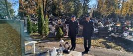 Na zdjęciu widać dwóch strażaków stojących przed grobem kolegi strażaka. Strażacy z stoją na baczność oddając hołd zmarłemu po uprzednim zapaleniu znicza. Jeden z nich ubrany jest w umundurowanie wyjściowe, drugi zaś w służbowe. Zdjęcie wykonane w porze południowej, mglistej. W tle nagrobki.