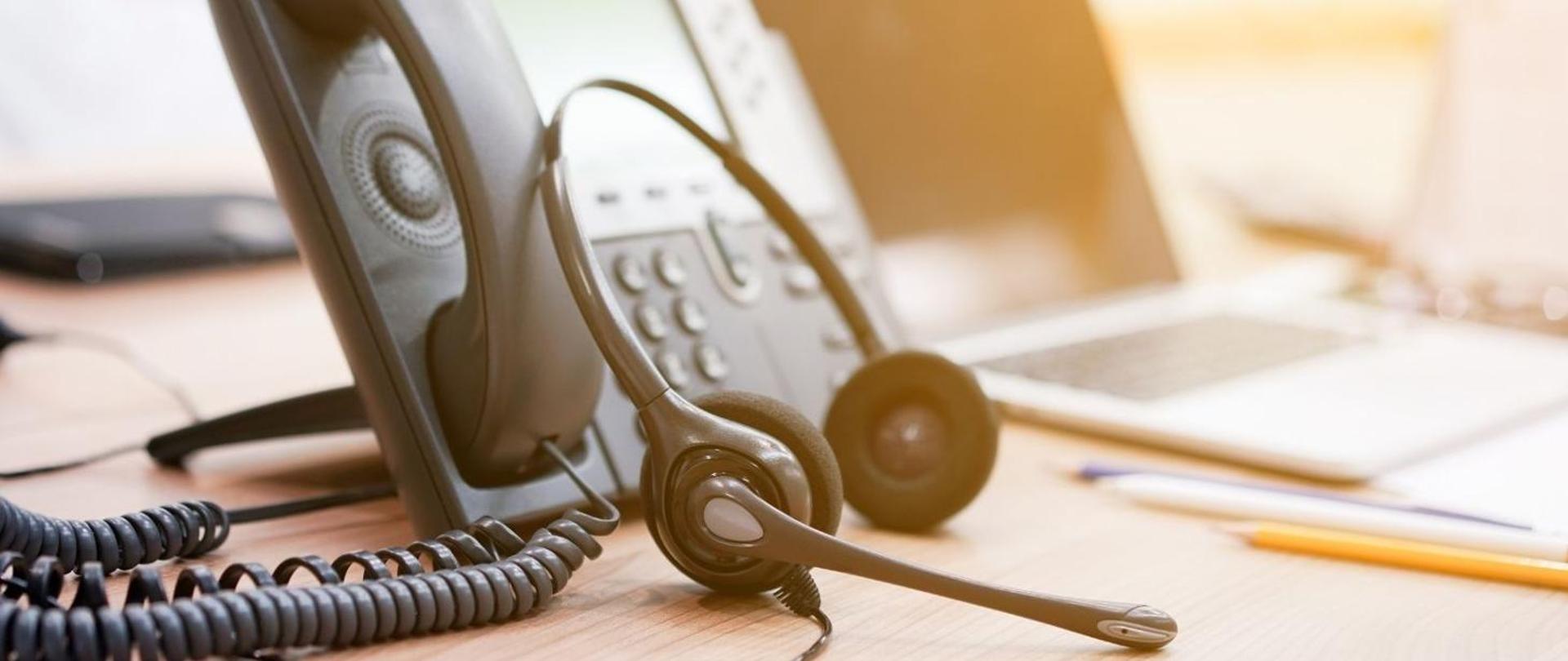 Telefon i słuchawki do obsługi infolinii. W tle otwarty laptop i przybory do pisania.