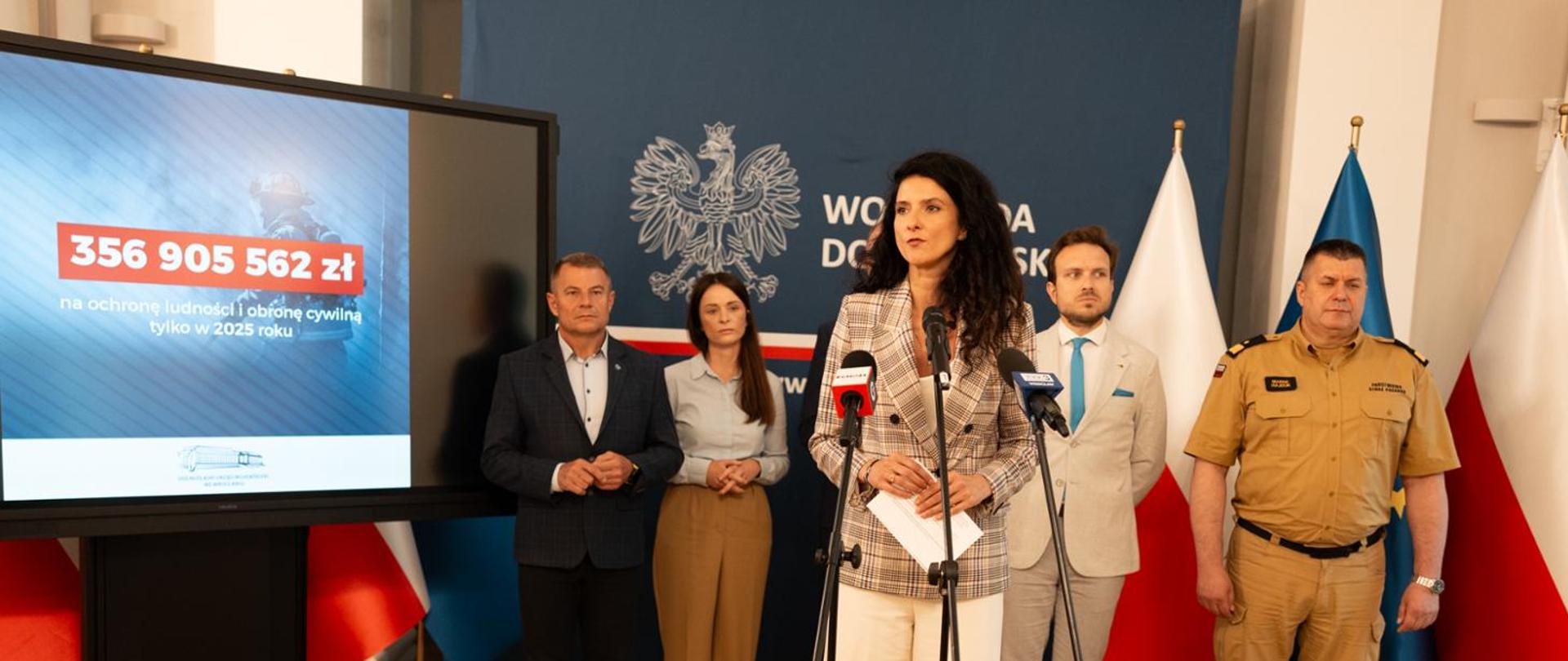 Konferencja prasowa wojewody Anny Żabskiej. Za wojewodą grupa ludzi, w tym strażak w mundurze, a także ekran z kwotą 356 905 562 zł 