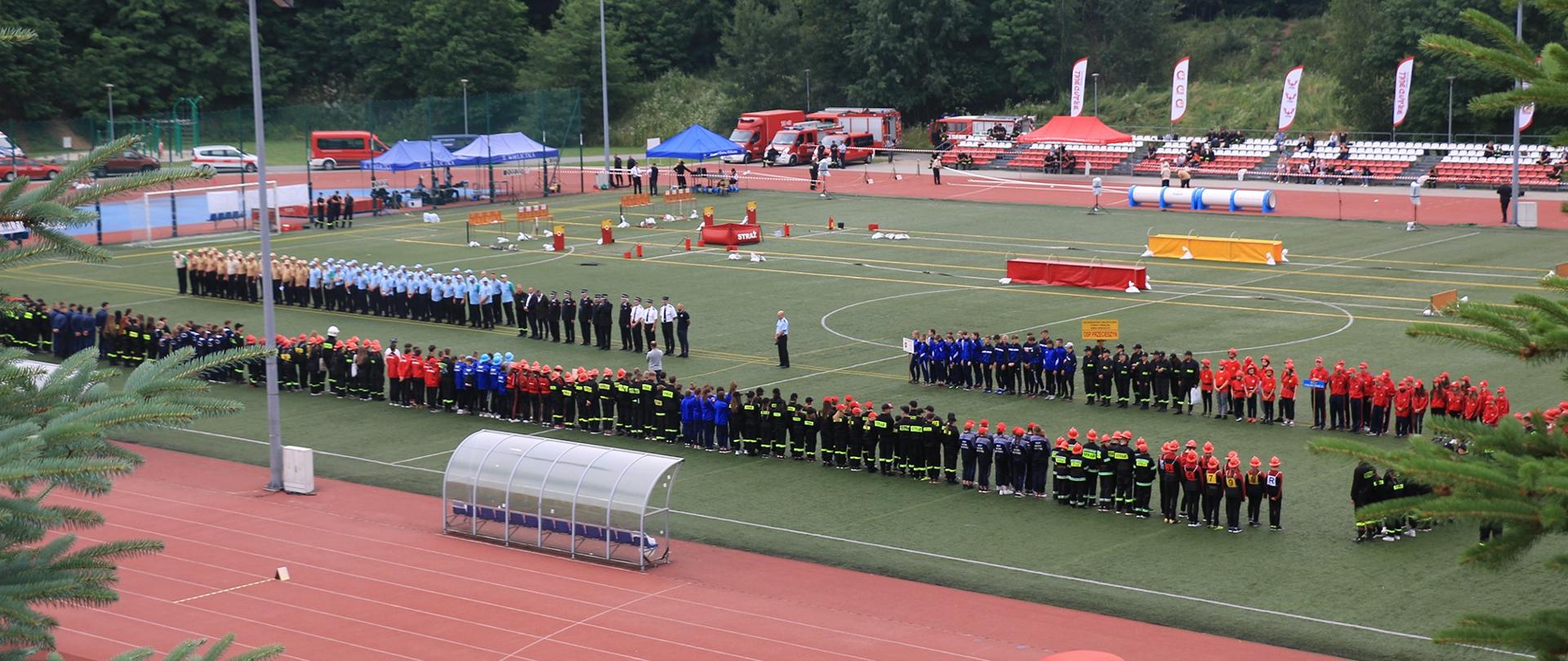 Boisko sportowe na którym ustawione w szeregu stoją drużyny przygotowane do zawadów.