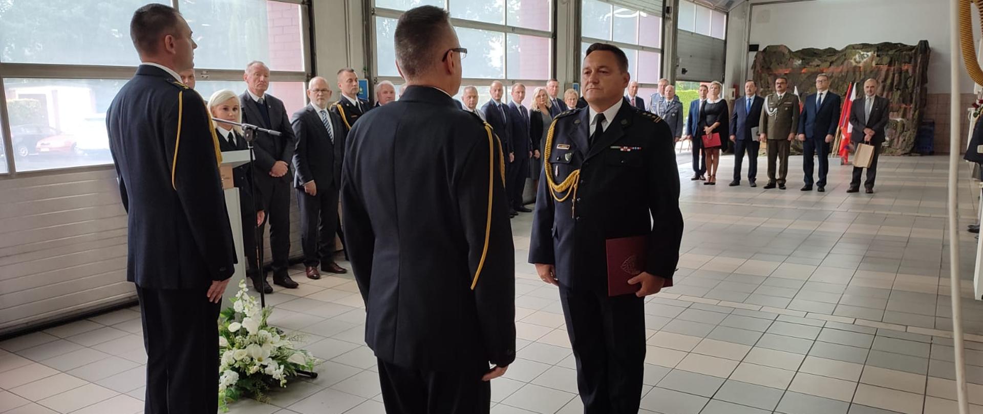 Strażacy w ubraniu wyjściowym stoją w garażu, z tył widać osoby w ubraniach cywilnych