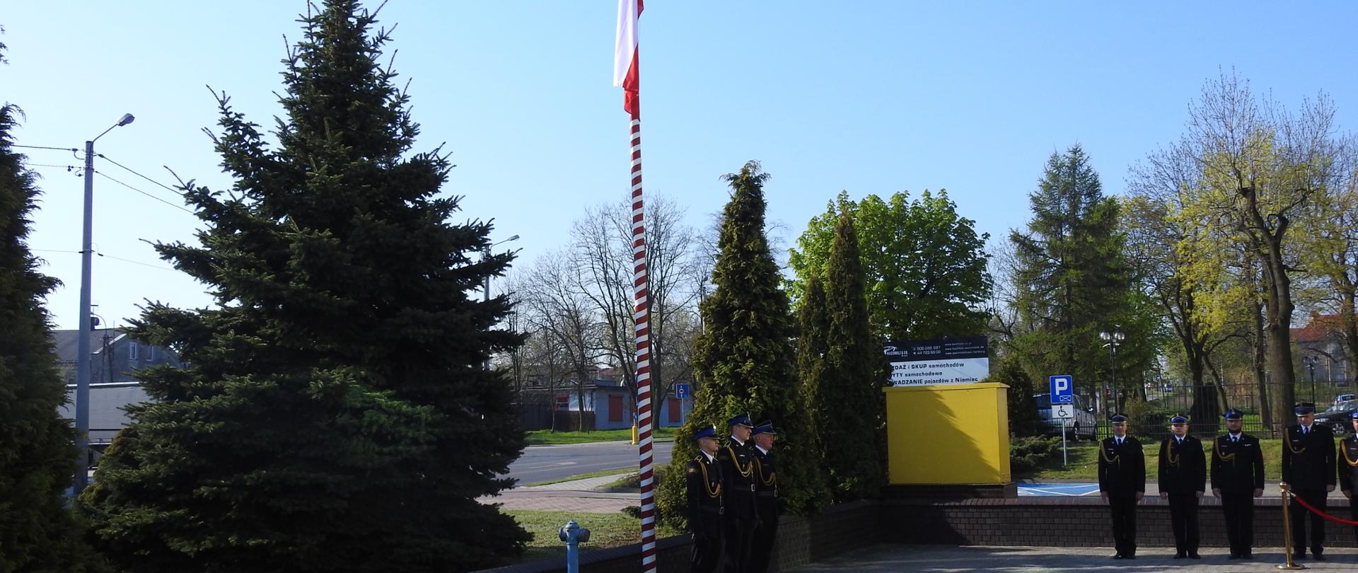 Na placu przy maszcie na którym powiewa biało-czerwona flaga stoi trzech strażaków. Na lewo od masztu stoi pięciu strażaków. Cały plac otoczony jest drzewami. 