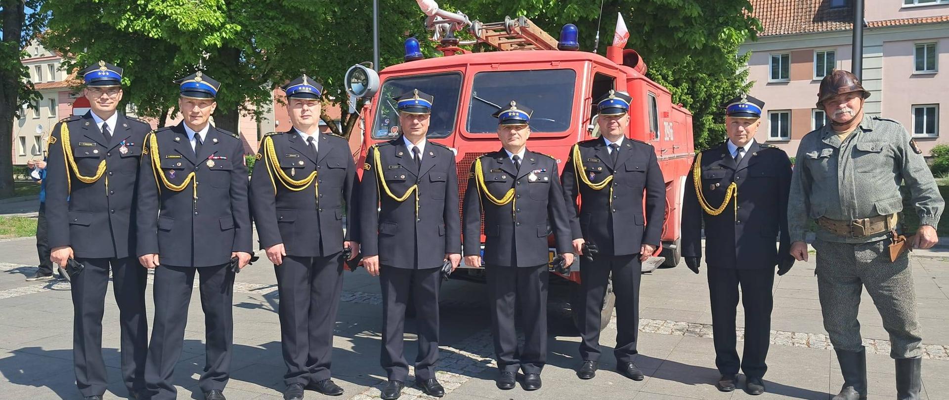 Na tle zabytkowego samochodu strażackiego stoi obok siebie grupa siedmiu strażaków w mundurach galowych. Z prawej strony na zdjęciu strażak ochotnik. Słonecznie.