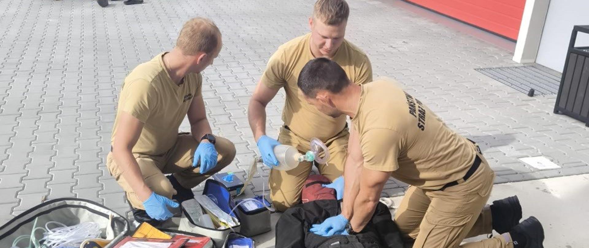 Zdjęcie przedstawia praktyczne ćwiczenia z zakresu kwalifikowanej pierwszej pomocy (KPP) prowadzone na zewnątrz jednostki Państwowej Straży Pożarnej.
Główne elementy na zdjęciu:
Strażacy w trakcie akcji: Trzech strażaków w piaskowych mundurach służbowych i niebieskich rękawiczkach jednorazowych prowadzi resuscytację manekina szkoleniowego ubranego w czarny kombinezon.
Jeden ze strażaków wykonuje uciski klatki piersiowej.
Drugi odpowiada za wentylację za pomocą worka samorozprężalnego (Ambu) z maską.
Trzeci asekuruje i przygotowuje sprzęt.
Sprzęt ratowniczy:
Otwarta, wyposażona torba medyczna R1 z zestawami do udrażniania dróg oddechowych i opatrunkami.
Pomarańczowa deska ortopedyczna ze stabilizatorami i pasami leżąca obok.
Tło i otoczenie:
Ćwiczenia odbywają się na placu manewrowym przed boksami garażowymi strażnicy (widoczne czerwone bramy).
W tle widać drugą grupę osób (prawdopodobnie ratowników medycznych lub innych uczestników) również prowadzącą ćwiczenia na podłożu.