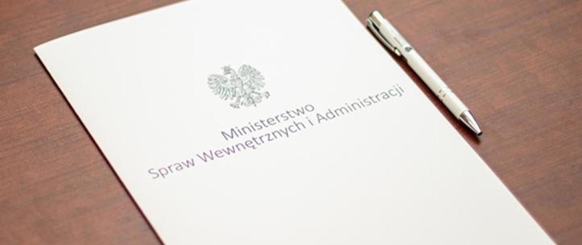 Zdjęcie przedstawiające białą teczkę z napisem Ministerstwo Spraw Wewnętrznych i Administracji leżącą na stole. Obok leżący biały długopis.