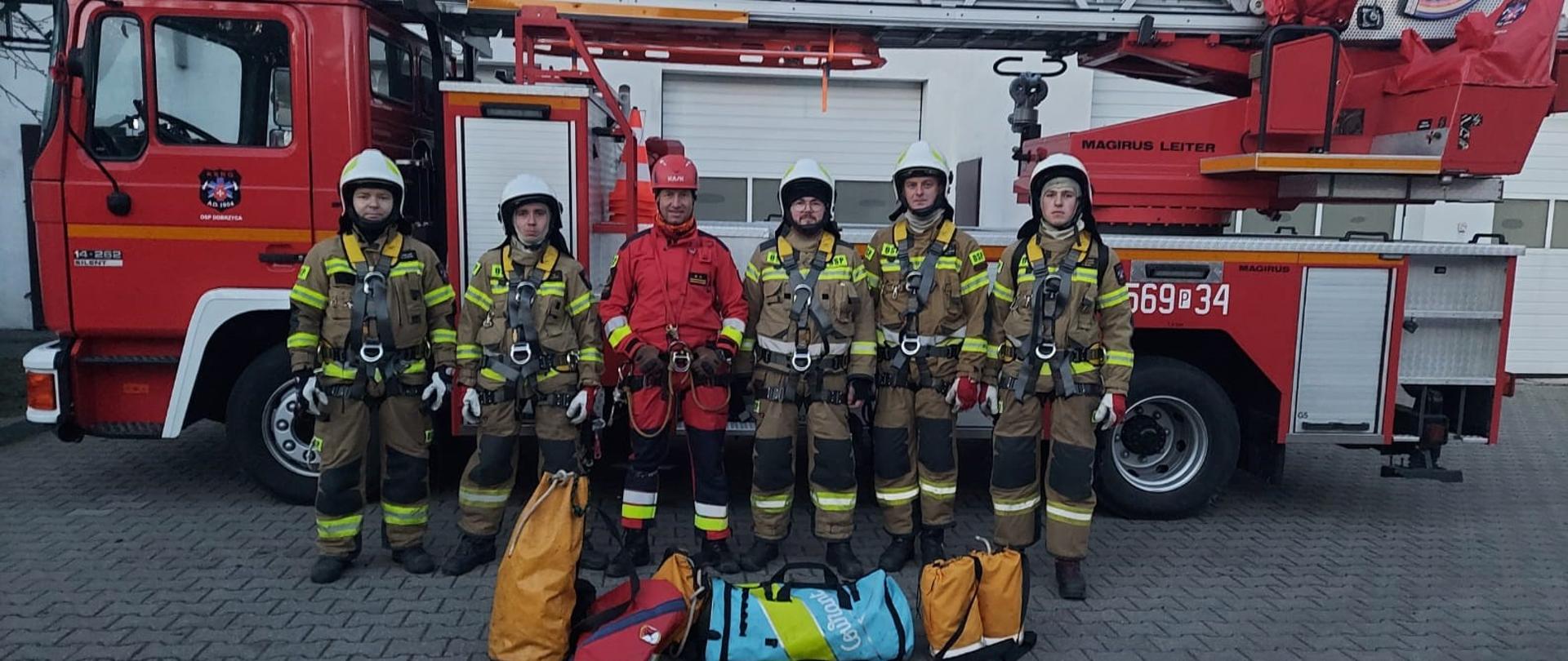 Sześciu strażaków stoi na tle drabiny strażackiej. ubrani w szelki ratownicze. Przed nimi stoją worki ze sprzętem do ratownictwa wysokościowego.