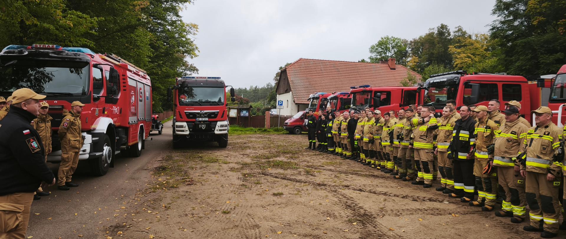 Grupa strażaków w mundurach stoi w szeregu na placu przed budynkiem. Wokół nich znajdują się czerwone wozy strażackie, a w tle widać drzewa i zabudowania.