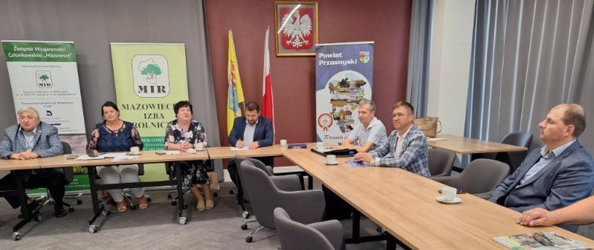 Zdjęcie przedstawia spotkanie w sali konferencyjnej – przy dwóch stołach w kształcie litery U siedzi kilkoro osób, na ścianie za nimi widoczne są banery Mazowieckiej Izby Rolniczej i Powiatu Przasnyskiego oraz godło Polski i flaga państwowa.