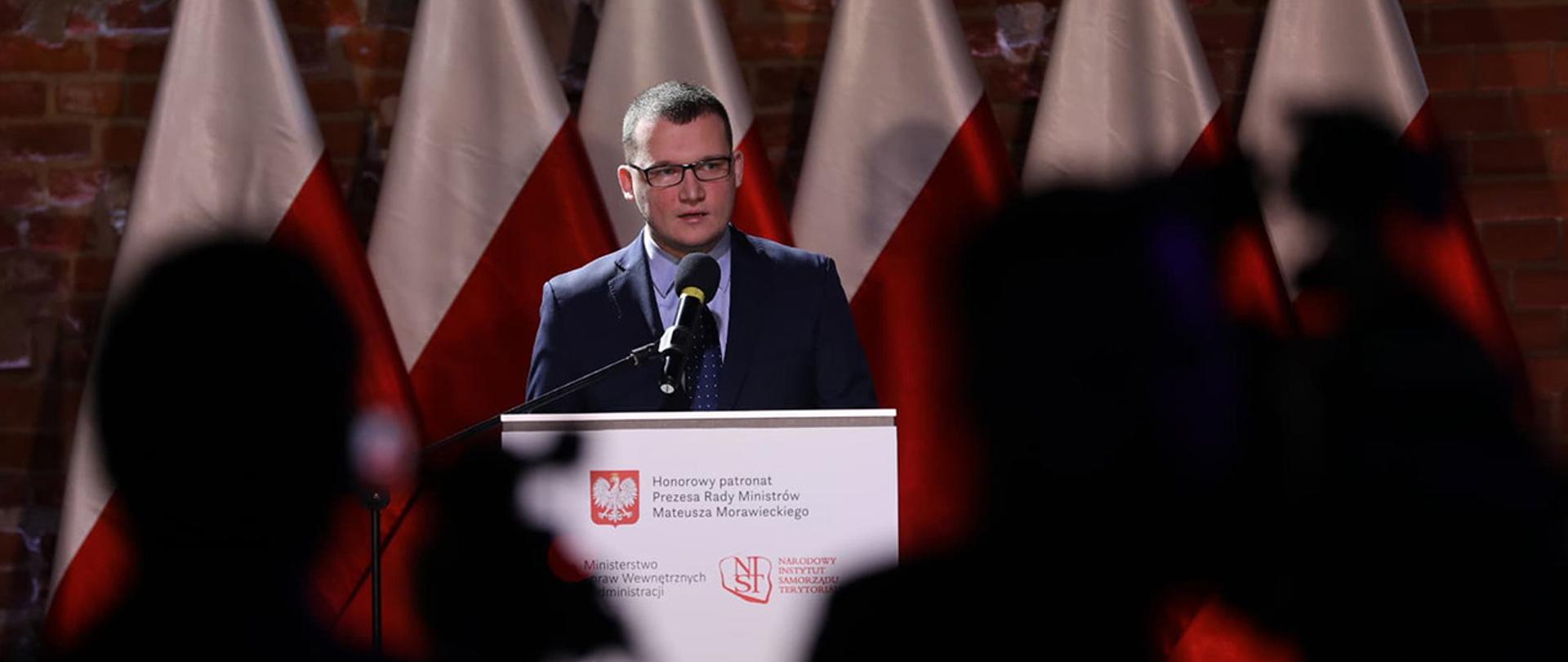 Na zdjęciu widać wiceministra Pawła Szefernakera stojącego przy mównicy i przemawiającego. W tle widać rząd flag Polski.